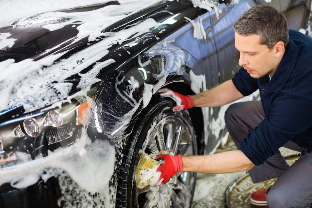 a detailer washing a dark blue car with a very soapy sponge, wearing red gloves