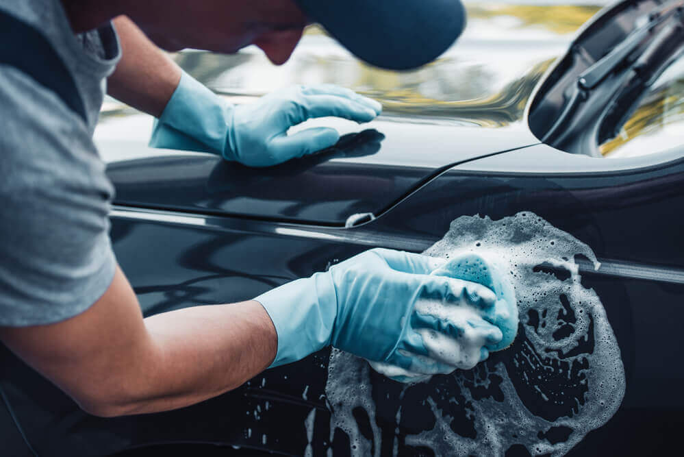 an over-the-shoulder view of a black car being washed with a soapy sponge